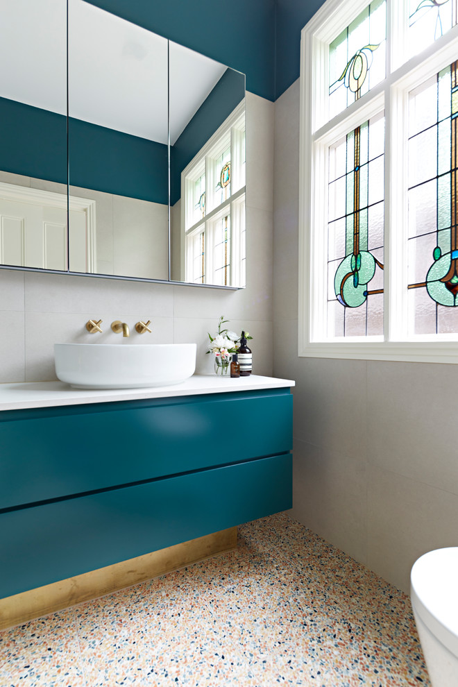 Inspiration for a small contemporary ensuite bathroom in Melbourne with green cabinets, a shower/bath combination, beige tiles, terrazzo flooring, a hinged door, white worktops, freestanding cabinets, a built-in bath, a wall mounted toilet, stone tiles, beige walls, a vessel sink, engineered stone worktops and multi-coloured floors.