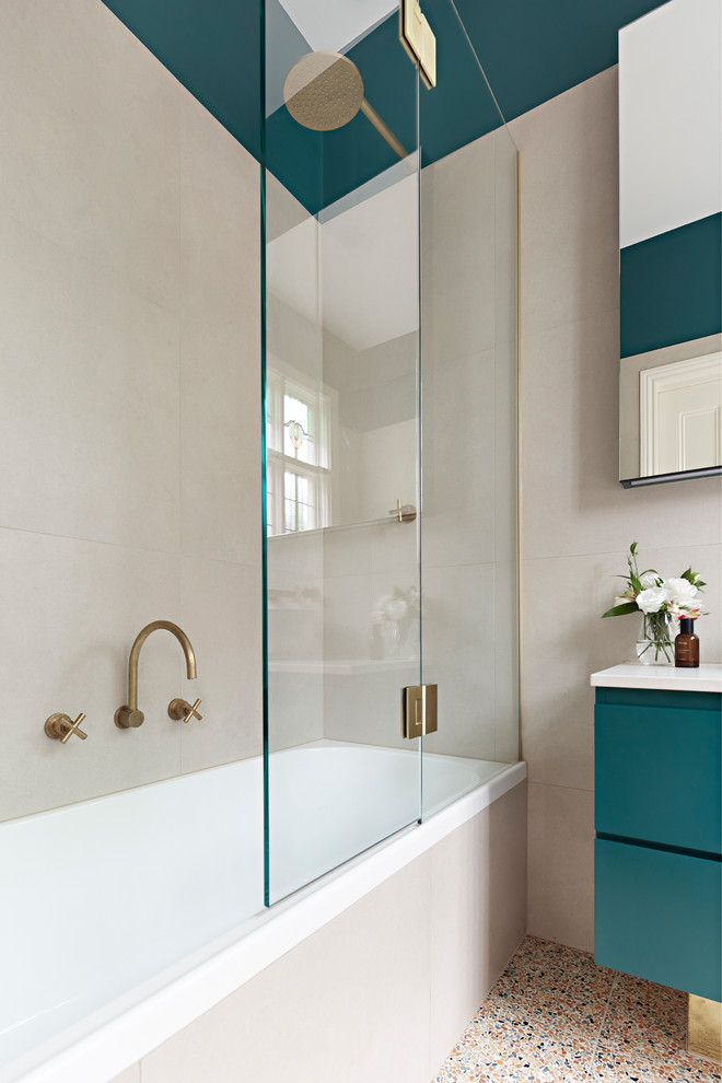 Photo of a small contemporary ensuite bathroom in Melbourne with green cabinets, a shower/bath combination, beige tiles, terrazzo flooring, a hinged door, white worktops, freestanding cabinets, a built-in bath, a wall mounted toilet, stone tiles, beige walls, a vessel sink, engineered stone worktops and multi-coloured floors.