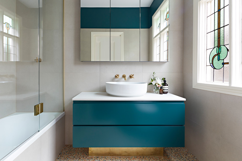 This is an example of a small contemporary ensuite bathroom in Melbourne with green cabinets, a shower/bath combination, beige tiles, terrazzo flooring, a hinged door, white worktops, freestanding cabinets, a built-in bath, a wall mounted toilet, stone tiles, beige walls, a vessel sink, engineered stone worktops and multi-coloured floors.