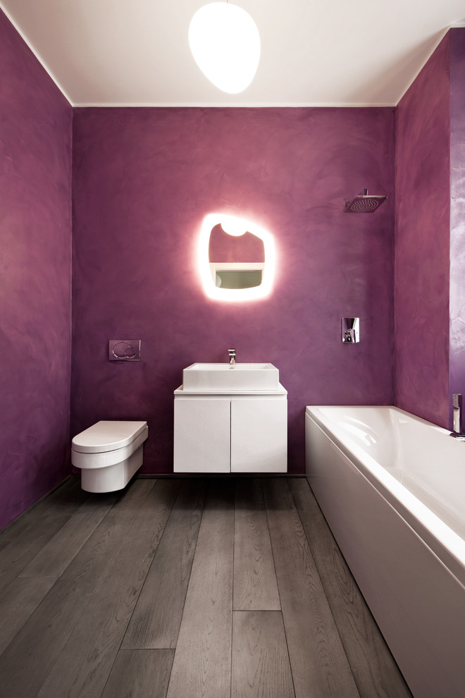 Diseño de cuarto de baño contemporáneo con lavabo sobreencimera, armarios con paneles lisos, puertas de armario blancas, bañera encastrada, sanitario de pared, paredes púrpuras, suelo de madera oscura y espejo con luz