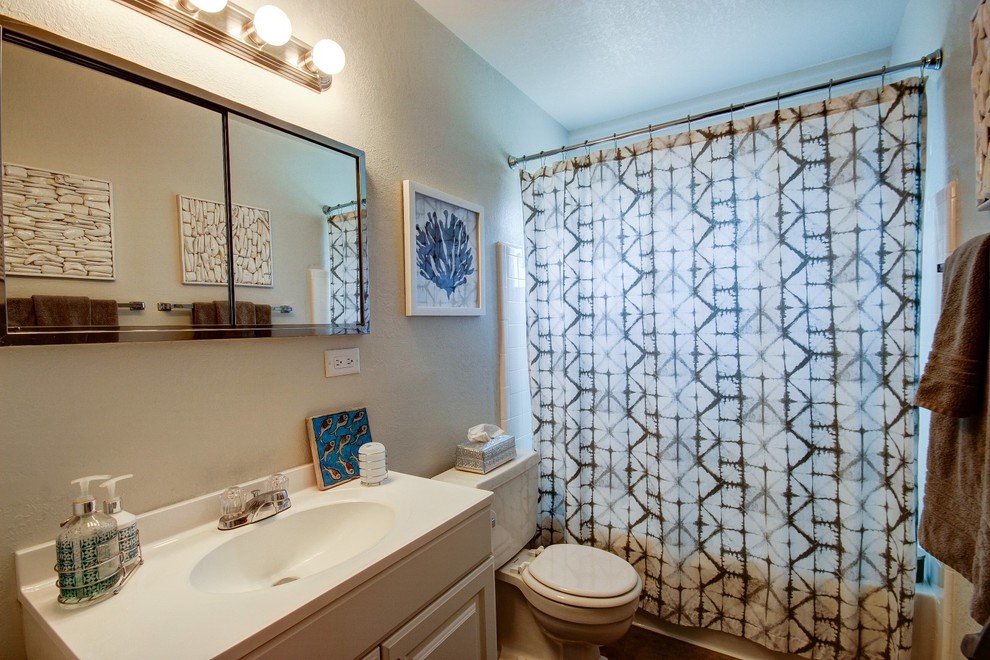 Photo of a small beach style ensuite bathroom in Minneapolis with an integrated sink, raised-panel cabinets, white cabinets, solid surface worktops, an alcove bath, a shower/bath combination, a two-piece toilet, white tiles, metro tiles, grey walls, concrete flooring and a shower curtain.