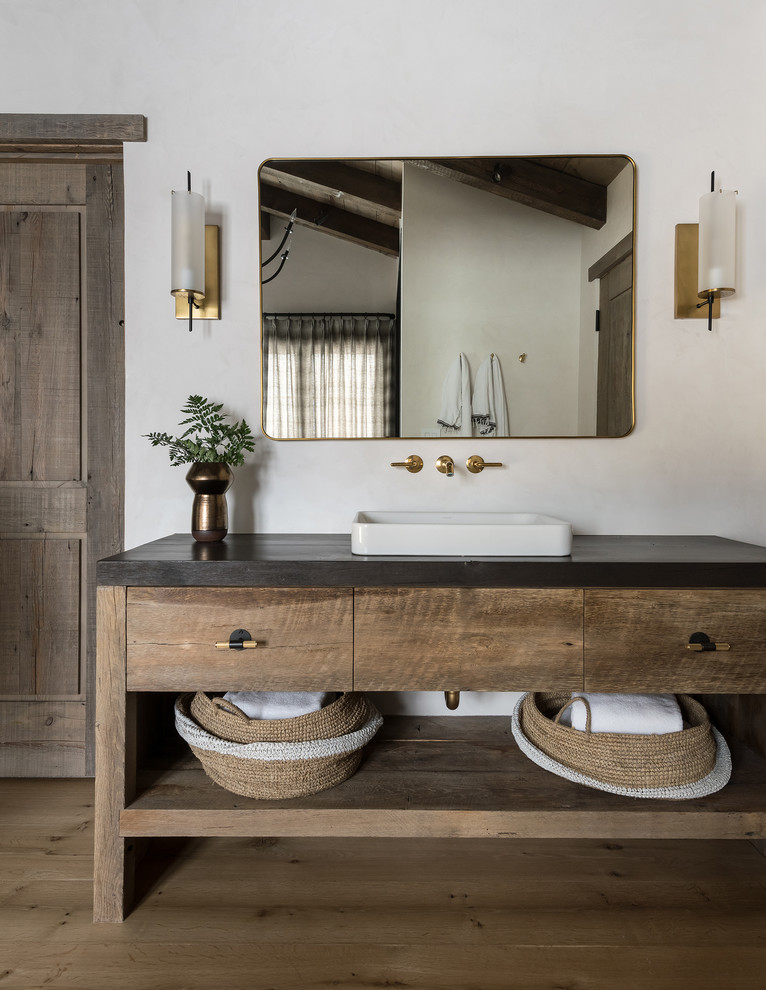 Diseño de cuarto de baño rural con puertas de armario de madera oscura, paredes blancas, suelo de madera en tonos medios, lavabo sobreencimera, suelo marrón, encimeras negras y armarios con paneles lisos