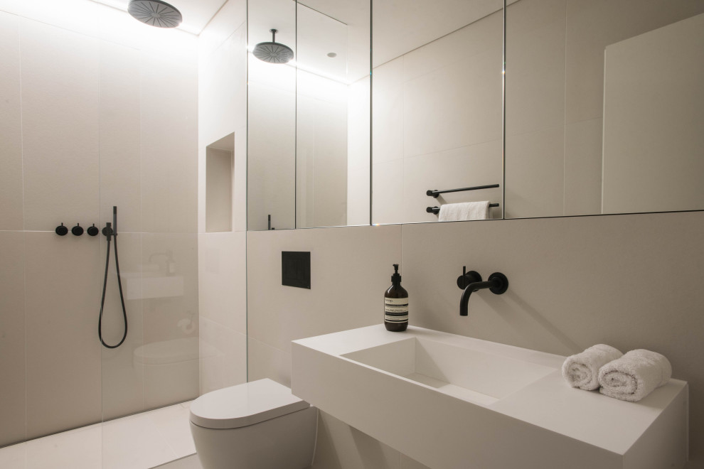 Idéer för mellanstora funkis vitt en-suite badrum, med släta luckor, vita skåp, en öppen dusch, en vägghängd toalettstol, beige kakel, porslinskakel, beige väggar, klinkergolv i porslin, ett konsol handfat, bänkskiva i akrylsten, beiget golv och med dusch som är öppen