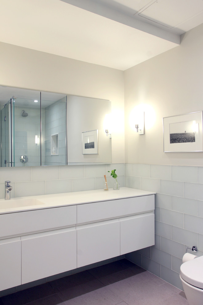 Foto på ett litet funkis en-suite badrum, med ett integrerad handfat, möbel-liknande, vita skåp, bänkskiva i akrylsten, en hörndusch, en vägghängd toalettstol, grå kakel, glaskakel, grå väggar och klinkergolv i porslin
