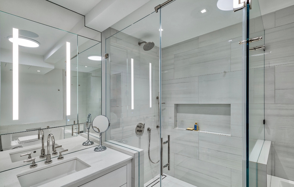 Corner shower - large contemporary master white tile and porcelain tile porcelain tile and white floor corner shower idea in New York with furniture-like cabinets, gray cabinets, a one-piece toilet, white walls, an undermount sink, marble countertops, a hinged shower door and white countertops