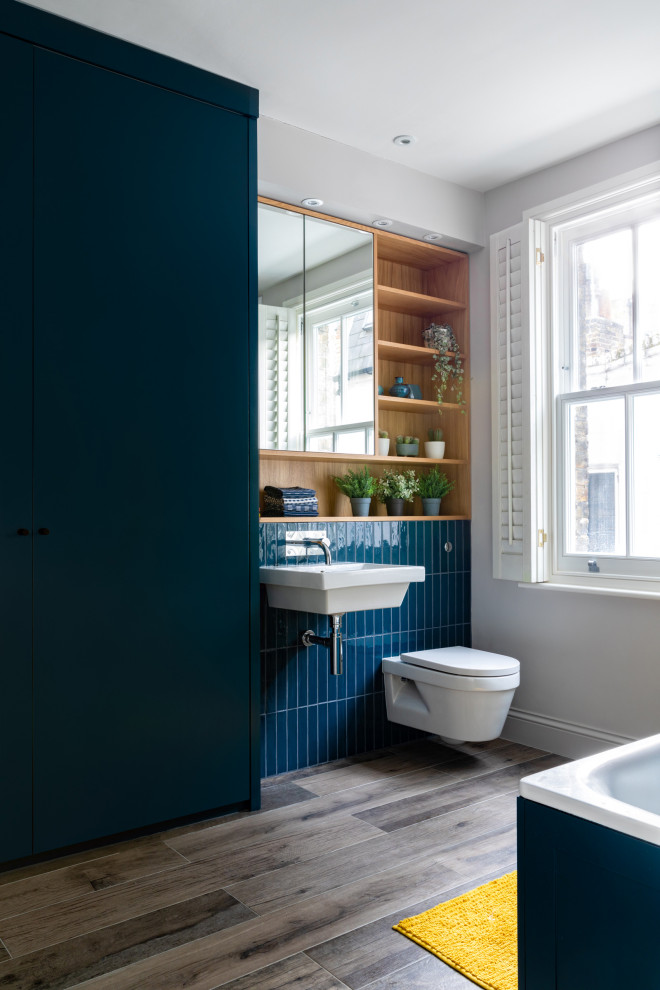 Inredning av ett klassiskt mellanstort blå blått badrum för barn, med släta luckor, blå skåp, ett platsbyggt badkar, en öppen dusch, en vägghängd toalettstol, blå kakel, keramikplattor, grå väggar, klinkergolv i porslin, ett väggmonterat handfat, träbänkskiva, grått golv och dusch med gångjärnsdörr