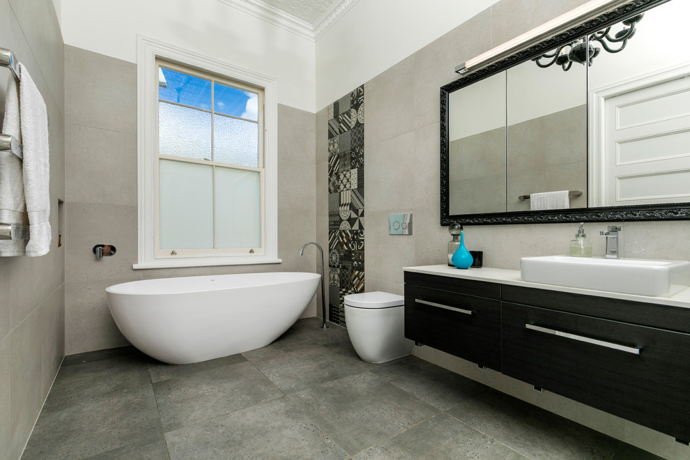 Inspiration for a large contemporary ensuite bathroom in Auckland with flat-panel cabinets, black cabinets, a freestanding bath, a wall mounted toilet, beige tiles, stone slabs, white walls, ceramic flooring, a vessel sink, engineered stone worktops and grey floors.