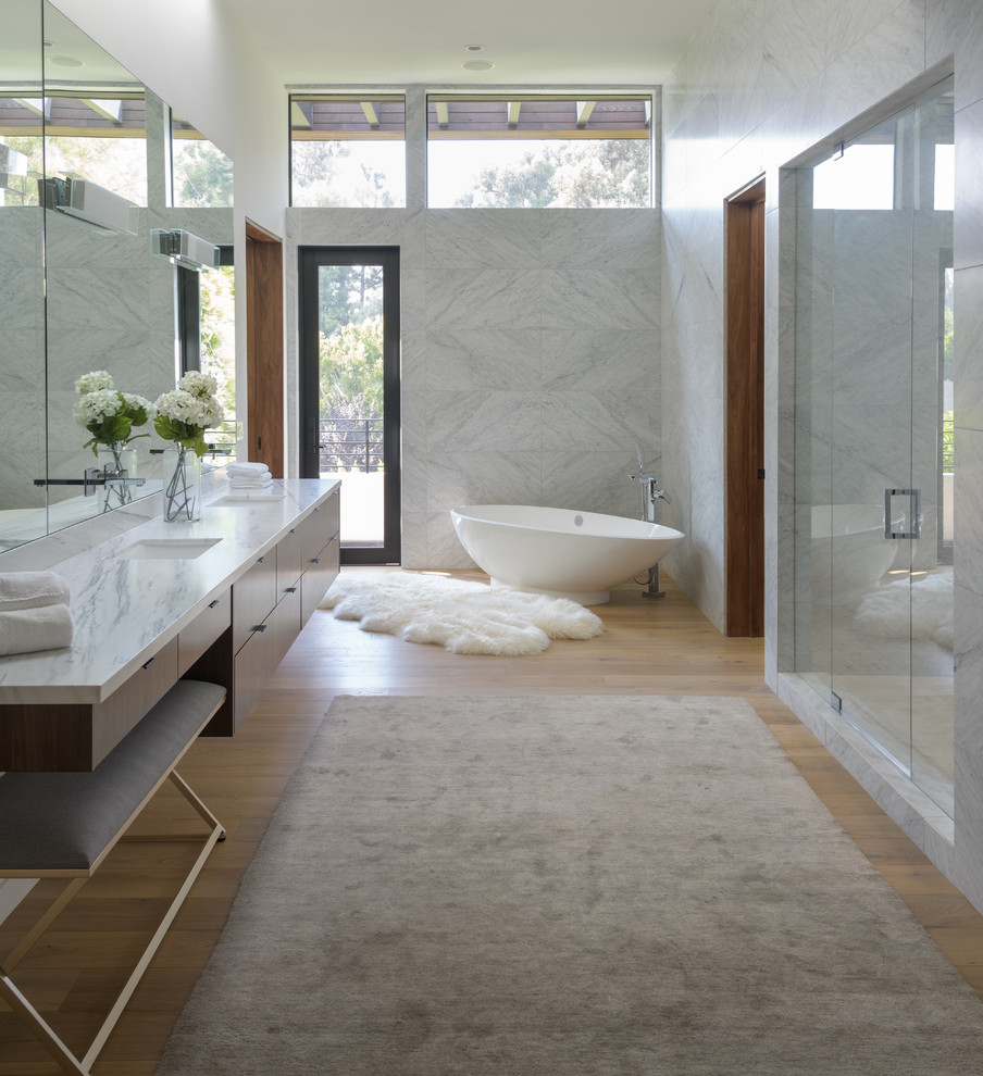 Inredning av ett modernt vit vitt badrum, med släta luckor, skåp i mörkt trä, ett fristående badkar, en dusch i en alkov, grå kakel, vita väggar, mellanmörkt trägolv, ett undermonterad handfat, brunt golv och dusch med gångjärnsdörr