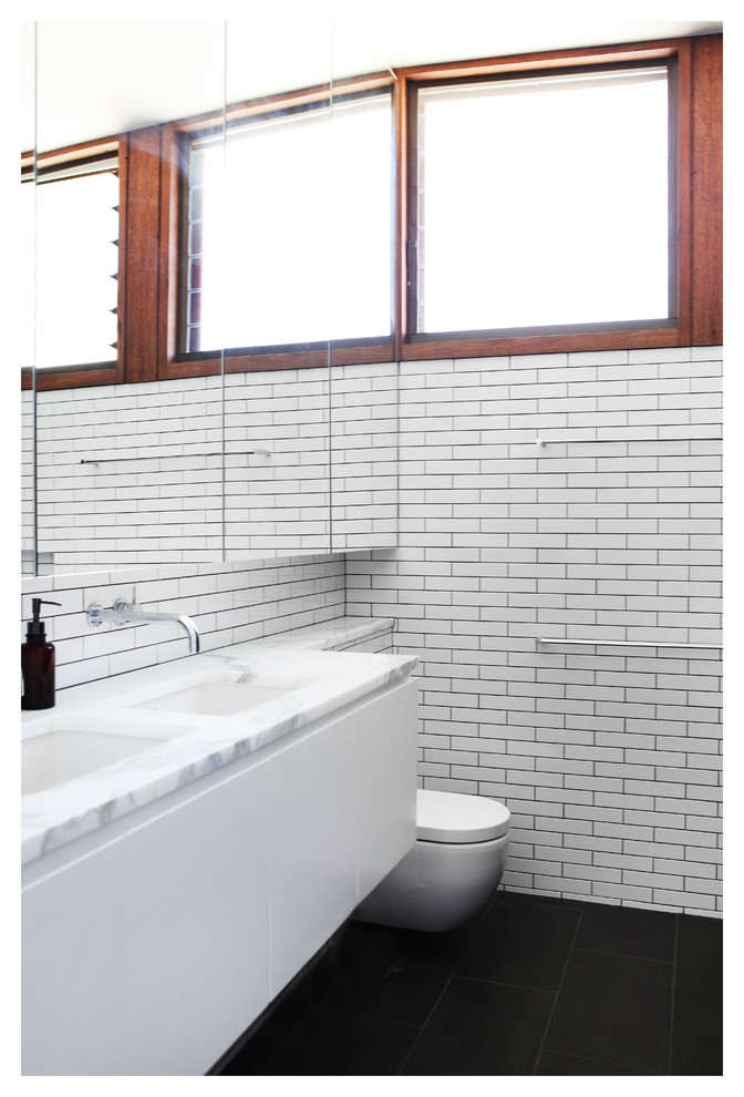 Inredning av ett modernt mellanstort badrum, med släta luckor, vita skåp, en toalettstol med hel cisternkåpa, vit kakel, tunnelbanekakel, klinkergolv i keramik, ett nedsänkt handfat, marmorbänkskiva och svart golv