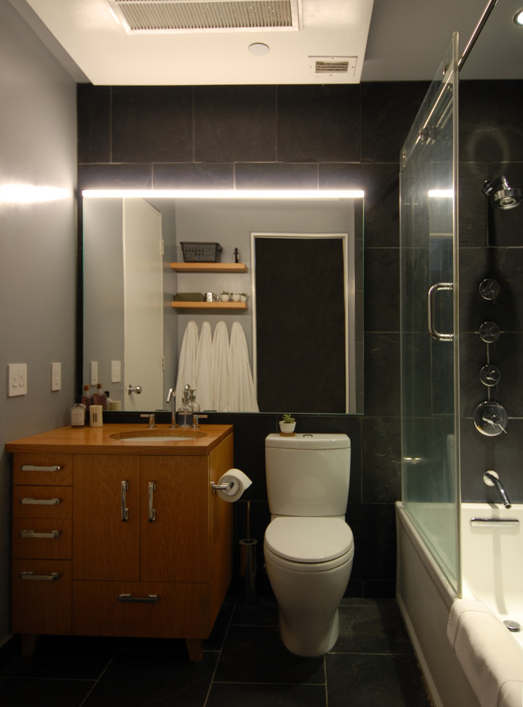 Свежая идея для дизайна: маленькая ванная комната в современном стиле с плоскими фасадами, светлыми деревянными фасадами, ванной в нише, душем над ванной, раздельным унитазом, черной плиткой, плиткой из сланца, черными стенами, полом из сланца, врезной раковиной, столешницей из дерева, черным полом, душем с раздвижными дверями и коричневой столешницей для на участке и в саду - отличное фото интерьера
