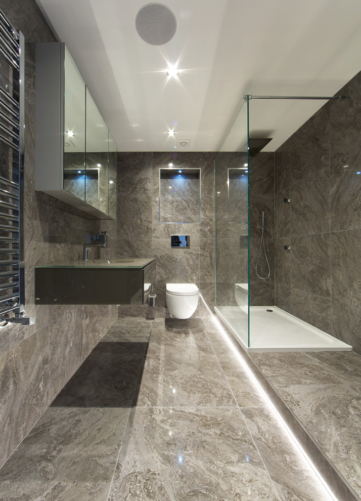 Modern inredning av ett mellanstort badrum med dusch, med luckor med glaspanel, grå skåp, en öppen dusch, en vägghängd toalettstol, grå kakel, stenkakel, grå väggar, klinkergolv i keramik, ett väggmonterat handfat och bänkskiva i glas