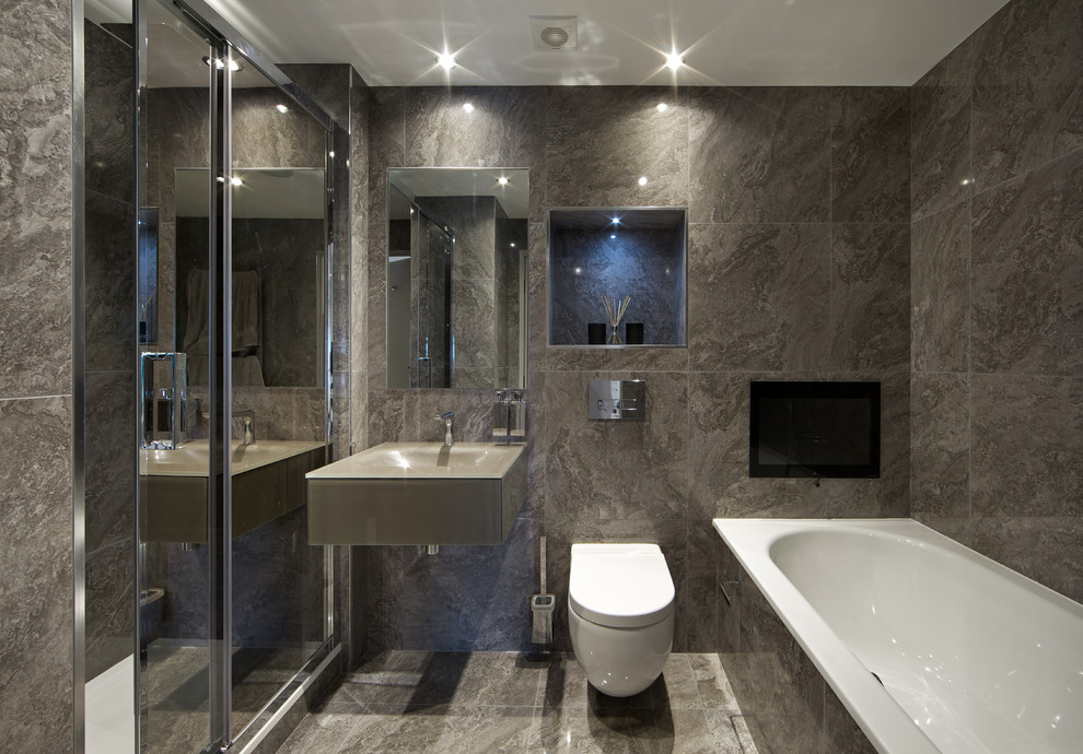 Exemple d'une douche en alcôve principale tendance de taille moyenne avec un placard à porte vitrée, des portes de placard grises, une baignoire posée, WC suspendus, un carrelage gris, un carrelage de pierre, un mur gris, un sol en carrelage de céramique, un lavabo suspendu et un plan de toilette en verre.