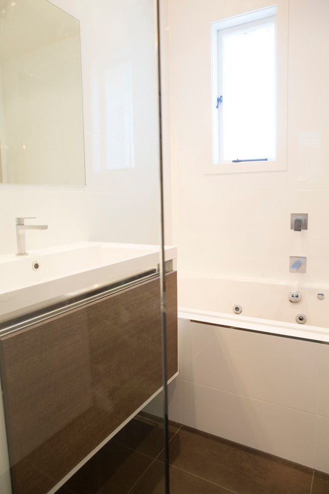 This is an example of a medium sized contemporary family bathroom in Auckland with flat-panel cabinets, medium wood cabinets, a built-in bath, a walk-in shower, a wall mounted toilet, brown tiles, ceramic tiles, white walls, ceramic flooring, an integrated sink and solid surface worktops.