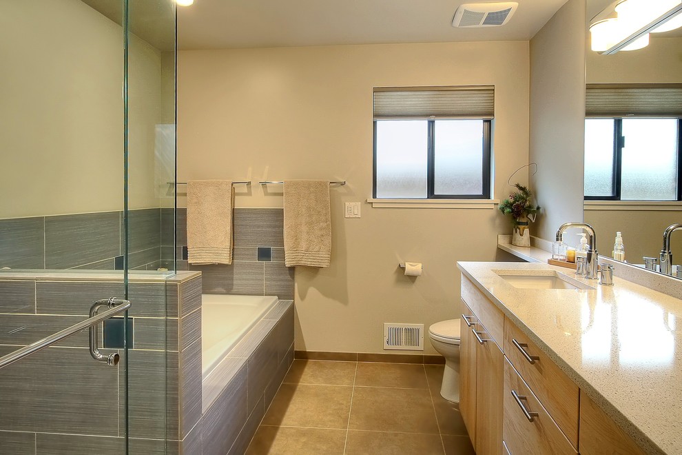 Diseño de cuarto de baño actual con lavabo bajoencimera, armarios con paneles lisos, bañera encastrada, ducha esquinera, baldosas y/o azulejos grises y puertas de armario de madera clara