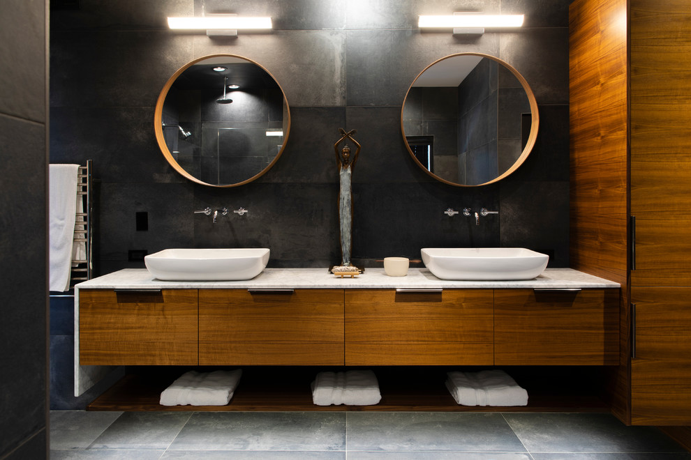 Cette photo montre une grande salle de bain principale tendance en bois brun avec un placard à porte plane, un carrelage noir, des carreaux de porcelaine, un mur noir, un sol en carrelage de porcelaine, une vasque, un plan de toilette en marbre, un sol gris et un plan de toilette blanc.