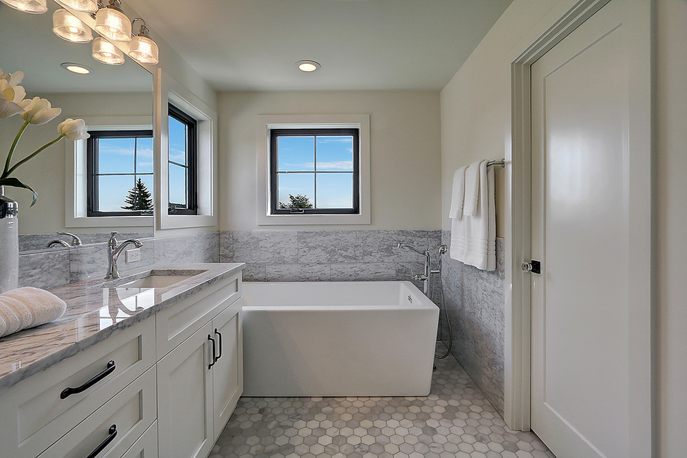 Inredning av ett klassiskt grå grått badrum, med luckor med infälld panel, vita skåp, ett fristående badkar, grå kakel, vita väggar, mosaikgolv, ett undermonterad handfat och vitt golv