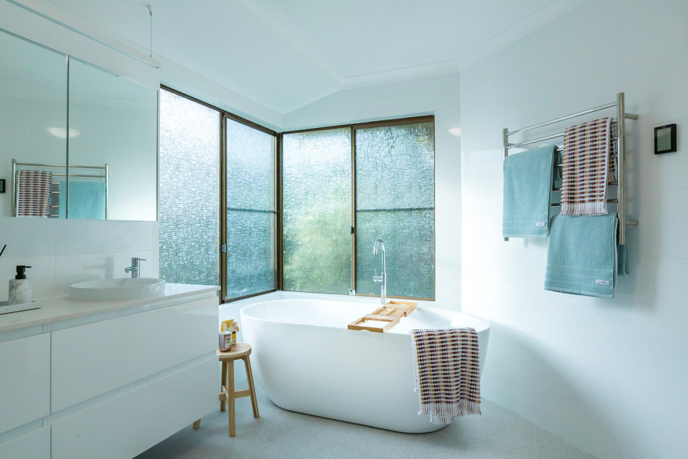 Стильный дизайн: большая главная ванная комната в стиле модернизм с плоскими фасадами, белыми фасадами, отдельно стоящей ванной, душем в нише, унитазом-моноблоком, белой плиткой, керамической плиткой, белыми стенами, полом из керамогранита, настольной раковиной, столешницей из искусственного кварца, разноцветным полом, душем с распашными дверями, белой столешницей, тумбой под две раковины и подвесной тумбой - последний тренд