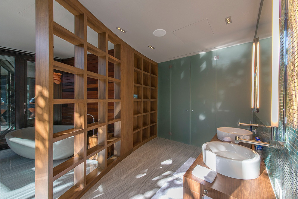 На фото: главная ванная комната среднего размера в современном стиле с плоскими фасадами, светлыми деревянными фасадами, отдельно стоящей ванной, инсталляцией, синей плиткой, стеклянной плиткой, настольной раковиной, столешницей из дерева и коричневой столешницей с