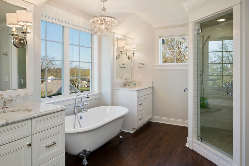 Inredning av ett klassiskt stort en-suite badrum, med ett badkar med tassar, luckor med infälld panel, vita skåp, en dusch i en alkov, vit kakel, stenkakel, vita väggar, mörkt trägolv, ett undermonterad handfat, marmorbänkskiva, brunt golv och dusch med gångjärnsdörr
