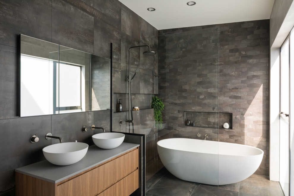 Modern inredning av ett grå grått badrum, med släta luckor, skåp i mellenmörkt trä, ett fristående badkar, grå kakel, ett fristående handfat och grått golv