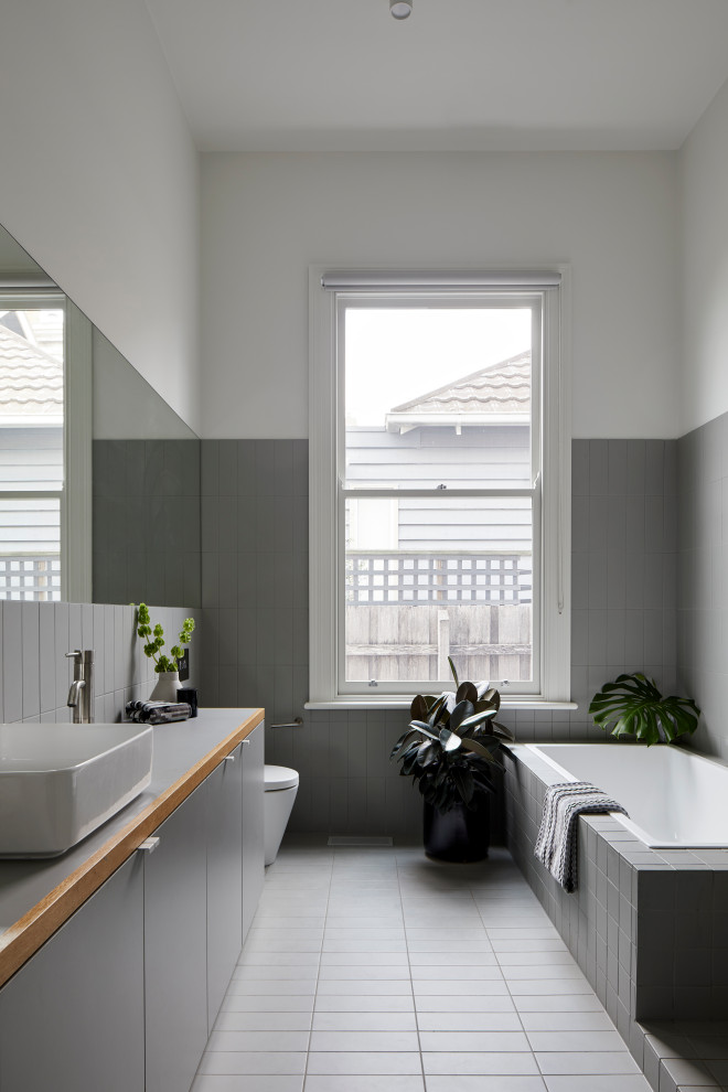 Cette photo montre une grande salle de bain principale tendance avec un placard à porte plane, des portes de placard grises, une baignoire d'angle, WC à poser, un carrelage gris, un mur blanc, un sol gris et un plan de toilette gris.