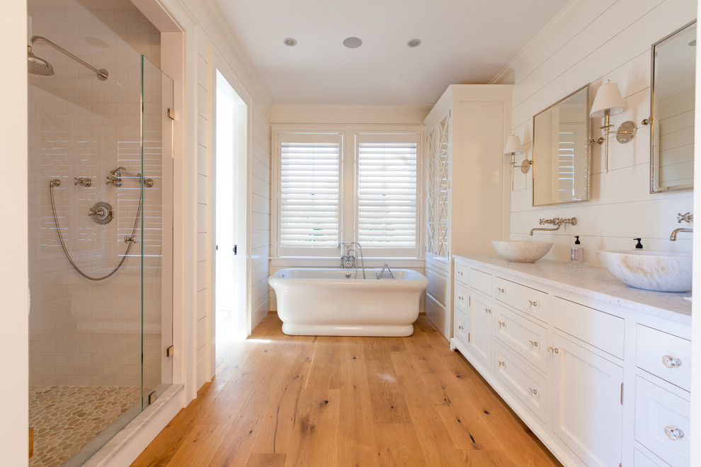 Exemple d'une grande douche en alcôve principale bord de mer avec une vasque, un placard avec porte à panneau encastré, des portes de placard blanches, un plan de toilette en marbre, une baignoire indépendante, un carrelage blanc, un carrelage métro, un mur blanc et un sol en bois brun.