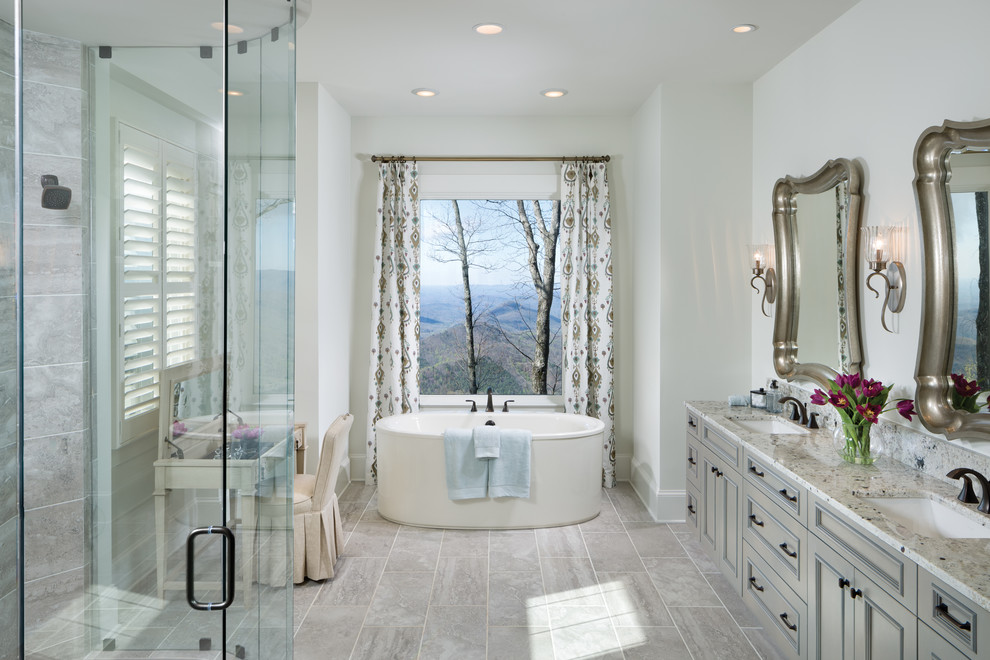 Cette image montre une salle de bain principale traditionnelle avec des portes de placard grises, une baignoire indépendante, un carrelage gris, des carreaux de céramique, un sol en carrelage de céramique, un lavabo encastré, un plan de toilette en granite, un mur blanc, un placard avec porte à panneau encastré, une cabine de douche à porte battante, une douche d'angle et un sol gris.