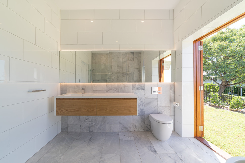This is an example of a large scandi family bathroom in Brisbane with beaded cabinets, light wood cabinets, a freestanding bath, a corner shower, a wall mounted toilet, white tiles, stone tiles, white walls, marble flooring, an integrated sink, solid surface worktops, grey floors and an open shower.