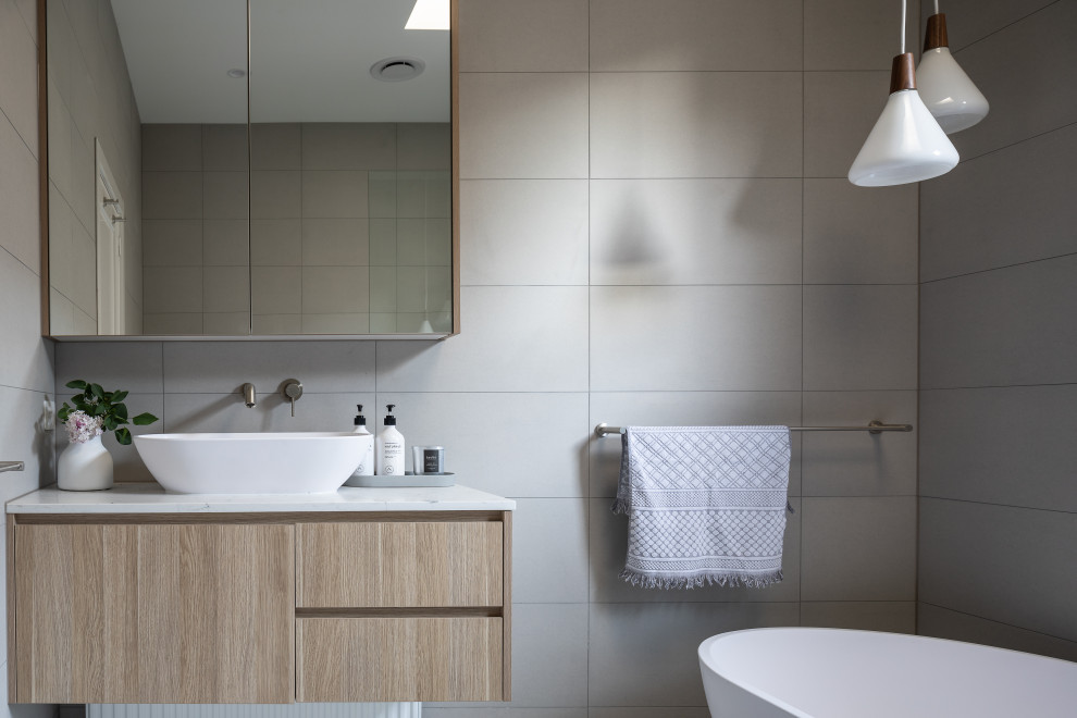 Modelo de cuarto de baño contemporáneo con armarios con paneles lisos, puertas de armario de madera clara, bañera exenta, baldosas y/o azulejos grises, lavabo sobreencimera y encimeras blancas