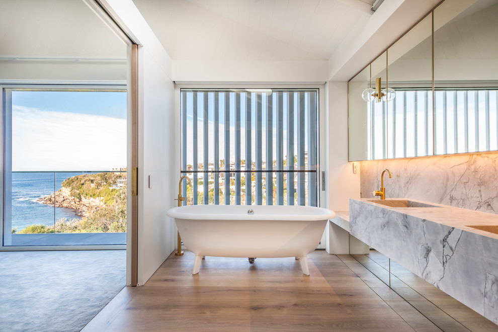 Modelo de cuarto de baño actual con bañera exenta, paredes blancas, suelo de madera en tonos medios, lavabo integrado, suelo marrón y encimeras grises