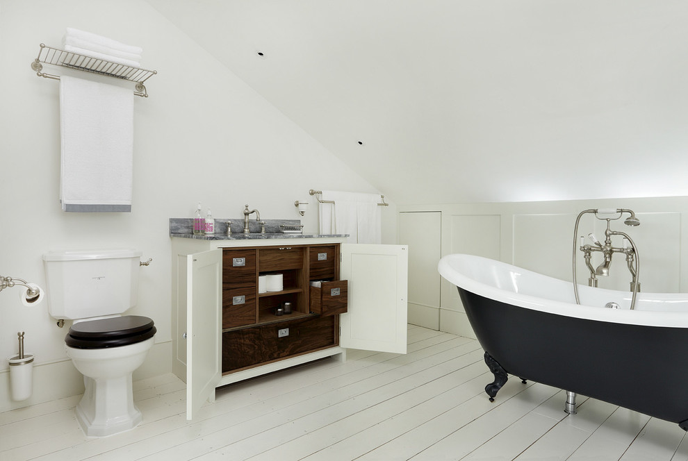 Klassisk inredning av ett en-suite badrum, med ett badkar med tassar, en toalettstol med separat cisternkåpa, vita väggar, målat trägolv och skåp i shakerstil