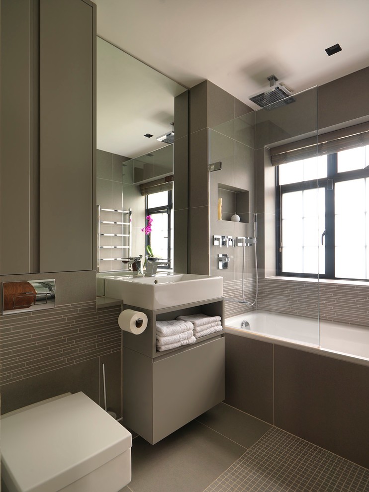 Modernes Badezimmer mit Waschtischkonsole, flächenbündigen Schrankfronten, grauen Schränken, Badewanne in Nische, Duschbadewanne, Wandtoilette und grauen Fliesen in London