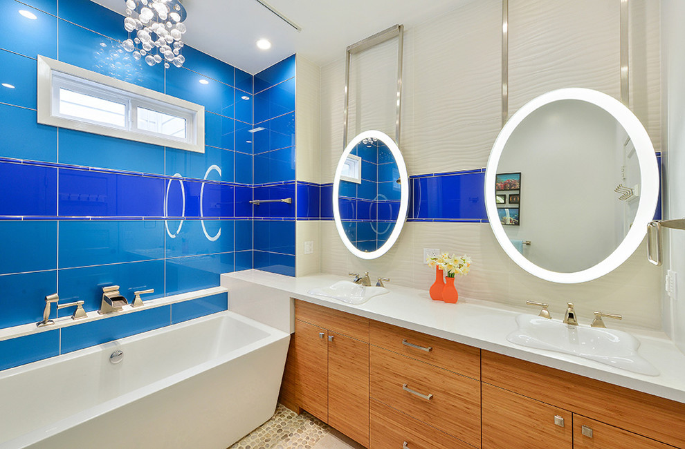 Cette photo montre une salle de bain principale tendance en bois clair de taille moyenne avec un placard à porte plane, un carrelage bleu, un carrelage en pâte de verre, un sol en galet, un lavabo intégré, un plan de toilette en surface solide, une baignoire indépendante et un mur multicolore.
