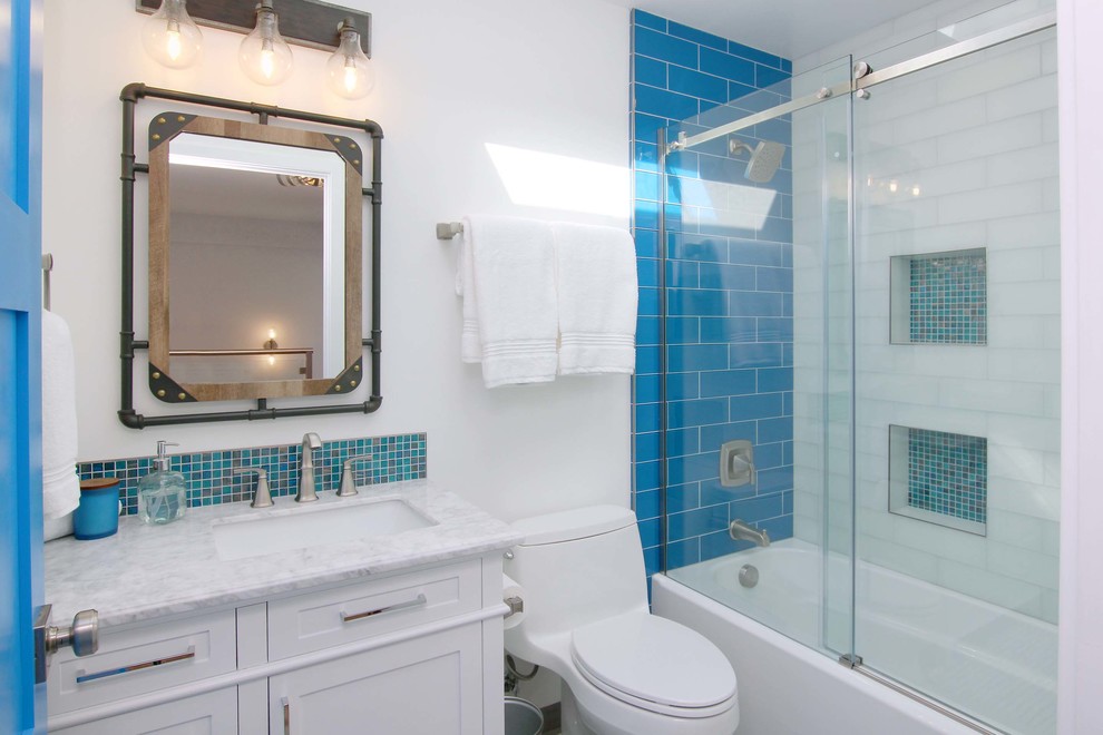 Cette image montre une salle d'eau marine de taille moyenne avec un placard à porte shaker, des portes de placard blanches, une baignoire en alcôve, un combiné douche/baignoire, WC à poser, un carrelage bleu, un carrelage blanc, un carrelage métro, un mur blanc, un lavabo encastré, un plan de toilette en marbre, une cabine de douche à porte coulissante et un plan de toilette gris.