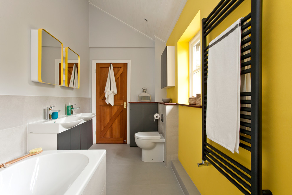 Diseño de cuarto de baño infantil actual con armarios con paneles lisos, puertas de armario grises, bañera encastrada, sanitario de una pieza, paredes amarillas y lavabo encastrado