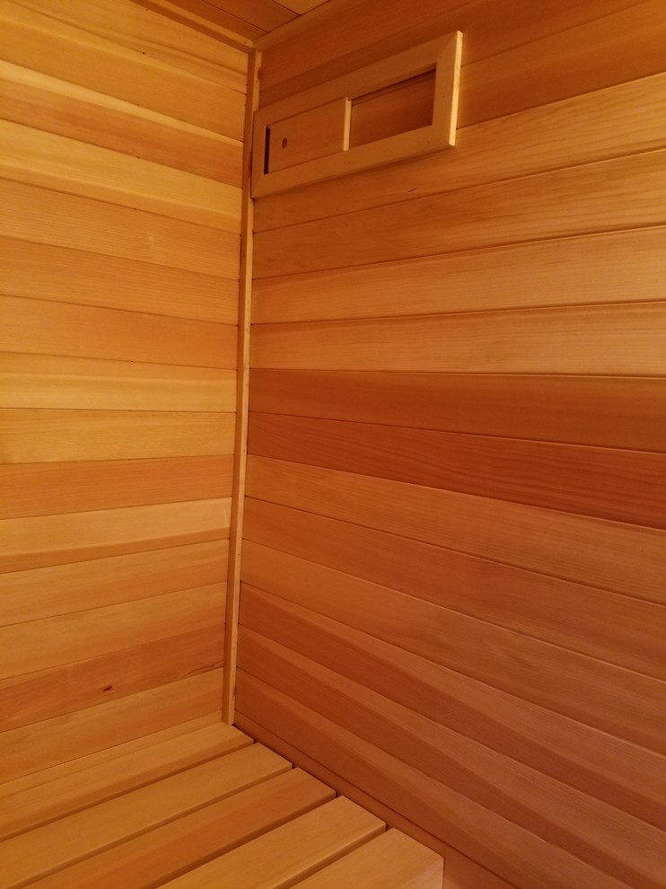Kleine Nordische Sauna mit profilierten Schrankfronten, hellbraunen Holzschränken, Eckdusche, Wandtoilette mit Spülkasten, beiger Wandfarbe, Travertin, Unterbauwaschbecken, Laminat-Waschtisch, buntem Boden und Falttür-Duschabtrennung in Grand Rapids