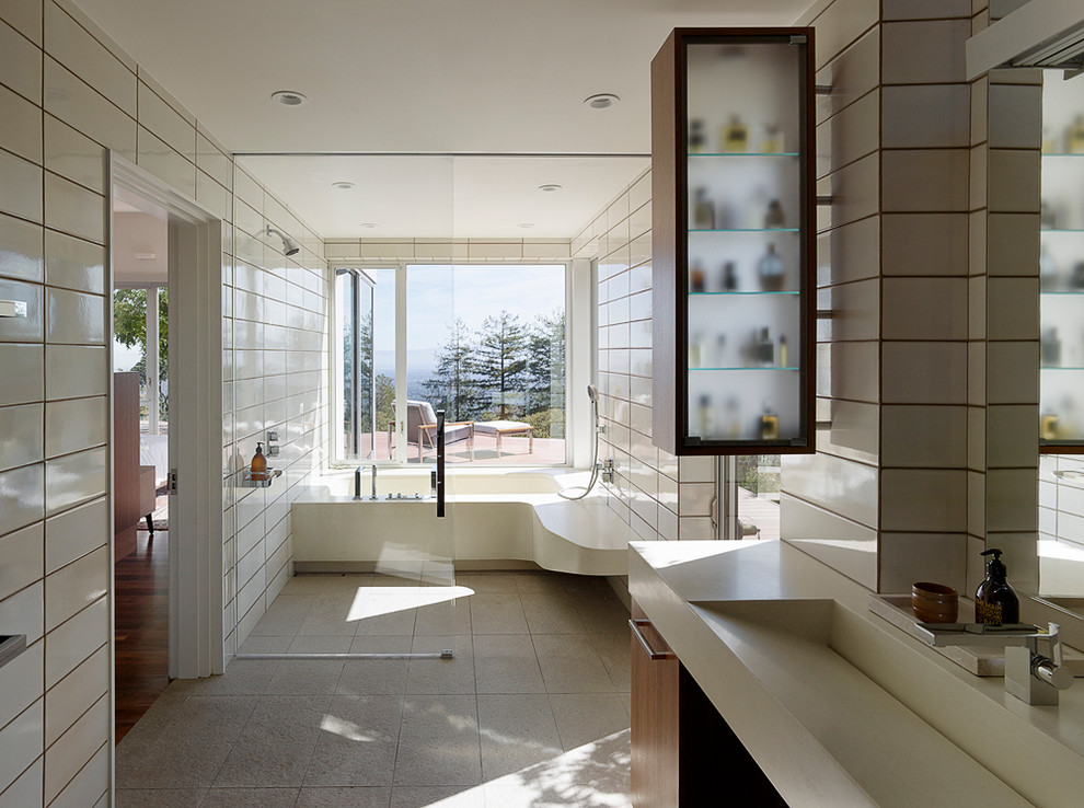 На фото: большая главная ванная комната в современном стиле с открытым душем, плоскими фасадами, фасадами цвета дерева среднего тона, ванной в нише, белой плиткой, керамогранитной плиткой, белыми стенами, бетонным полом, монолитной раковиной и серым полом с