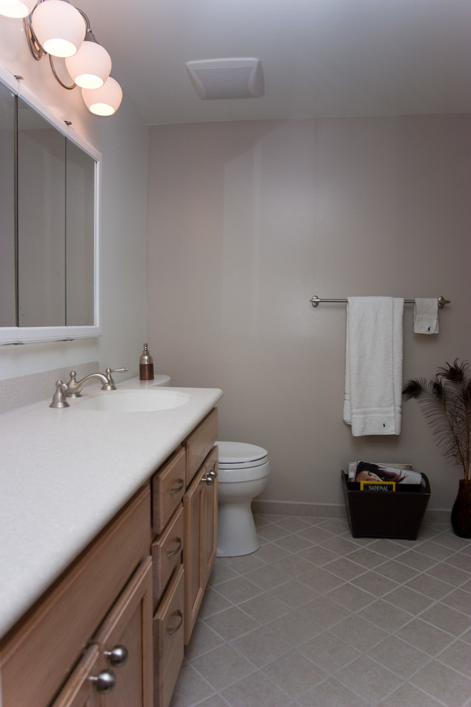 Klassisk inredning av ett litet en-suite badrum, med ett integrerad handfat, luckor med infälld panel, skåp i ljust trä, bänkskiva i akrylsten, ett badkar i en alkov, en dusch/badkar-kombination, en toalettstol med separat cisternkåpa, beige kakel, keramikplattor, beige väggar och klinkergolv i keramik
