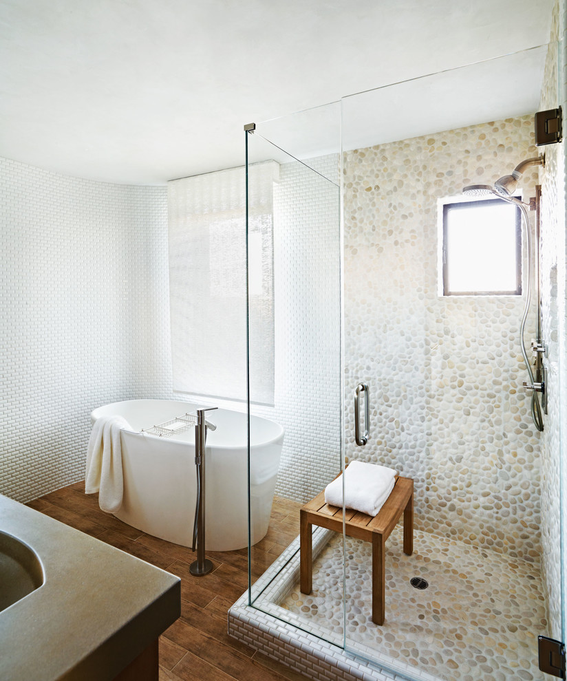 Cette image montre une salle de bain design avec une baignoire indépendante, une douche d'angle, un carrelage blanc, un mur blanc, une plaque de galets, un sol en galet et un banc de douche.
