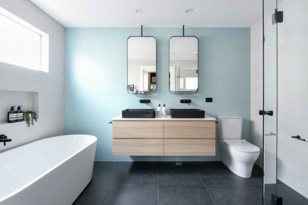 Cette image montre une salle de bain design en bois clair de taille moyenne avec un placard à porte plane, une baignoire indépendante, WC séparés, un carrelage bleu, un carrelage blanc, une vasque, un sol noir, un plan de toilette blanc, meuble double vasque et meuble-lavabo suspendu.