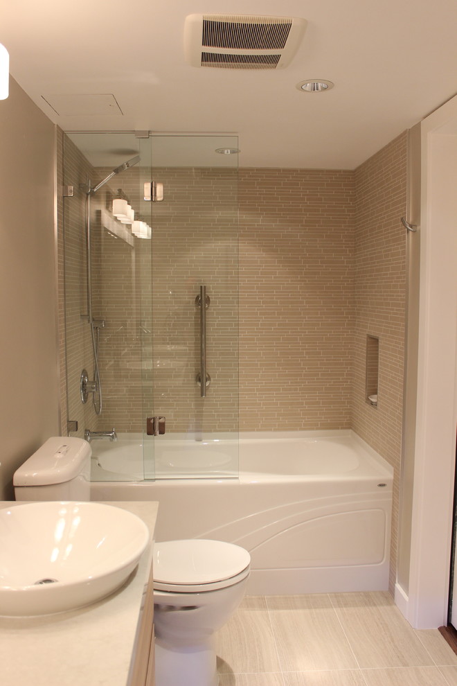 Klassisk inredning av ett badrum, med ett fristående handfat, släta luckor, skåp i ljust trä, bänkskiva i kvarts, ett badkar i en alkov, en dusch/badkar-kombination, en toalettstol med separat cisternkåpa och brun kakel
