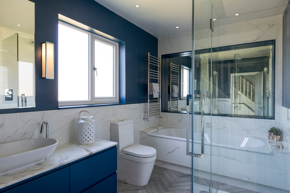 This is an example of a medium sized contemporary ensuite bathroom in London with flat-panel cabinets, blue cabinets, an alcove bath, a two-piece toilet, marble tiles, blue walls, a vessel sink, marble worktops, grey floors, white worktops and white tiles.