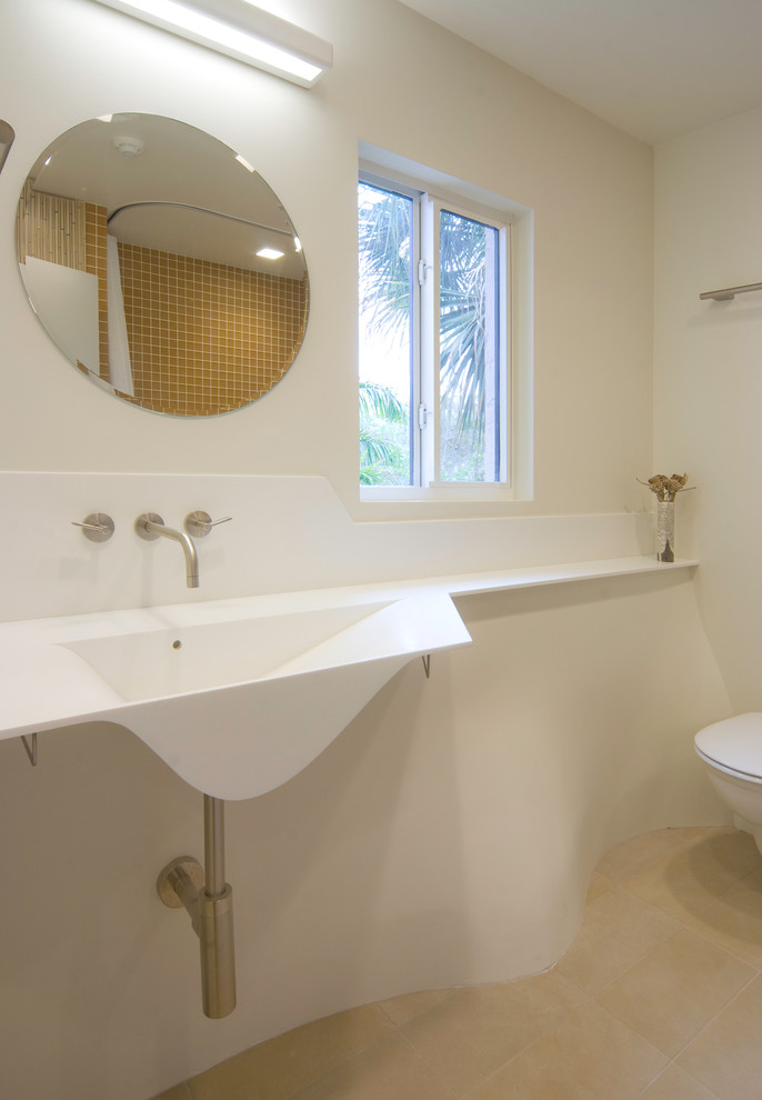 Example of a mid-sized trendy master orange tile and glass tile limestone floor and brown floor corner bathtub design in Tampa with flat-panel cabinets, light wood cabinets, a wall-mount toilet, beige walls, an integrated sink and solid surface countertops