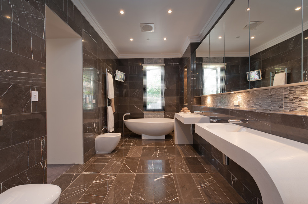 На фото: большая главная ванная комната в современном стиле с монолитной раковиной, мраморной столешницей, отдельно стоящей ванной, коричневой плиткой, керамогранитной плиткой, коричневыми стенами, полом из керамогранита и биде