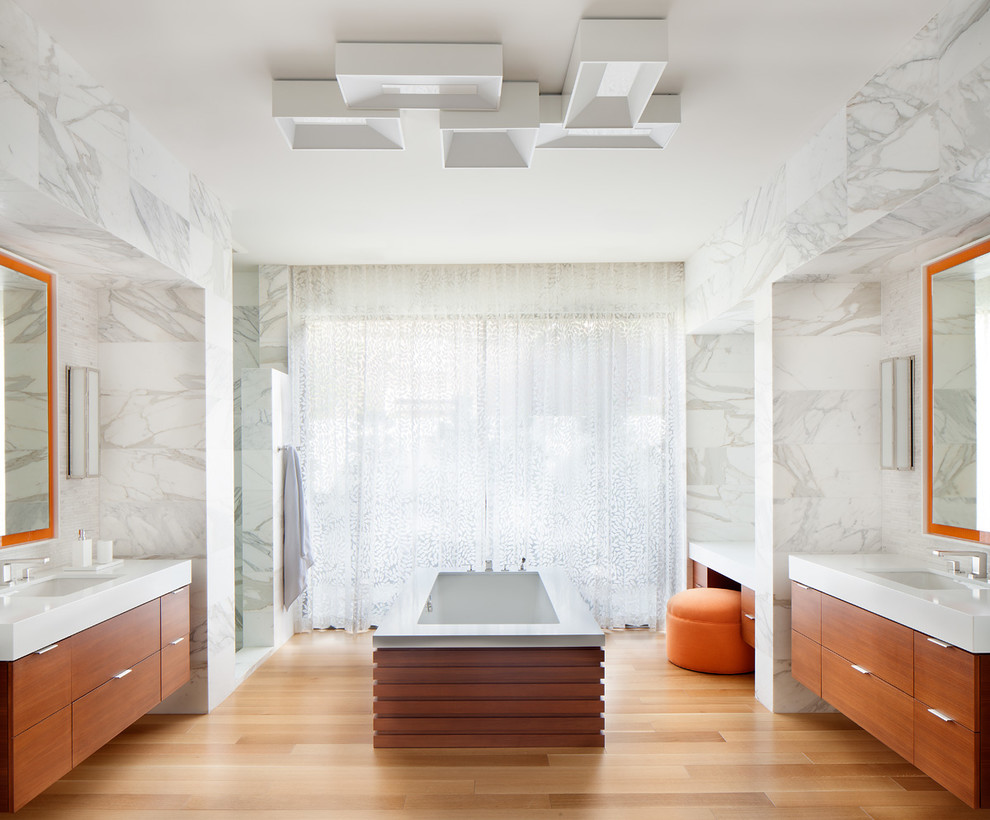Cette image montre une grande salle de bain principale design en bois brun avec un placard à porte plane, une baignoire encastrée, un carrelage blanc, un lavabo encastré, une douche d'angle, un carrelage de pierre, un mur blanc, parquet clair, un plan de toilette en surface solide et un sol beige.