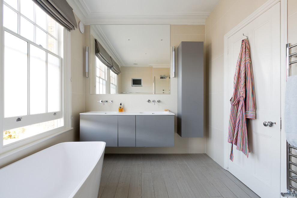 Modelo de cuarto de baño principal actual de tamaño medio con armarios con paneles lisos, puertas de armario grises, bañera exenta, paredes beige y suelo de madera en tonos medios