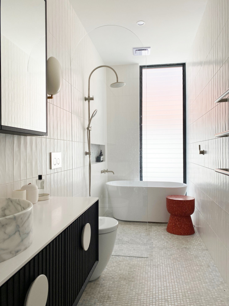 Imagen de cuarto de baño actual con puertas de armario negras, bañera exenta, baldosas y/o azulejos blancos, suelo con mosaicos de baldosas, lavabo sobreencimera, suelo blanco y encimeras blancas