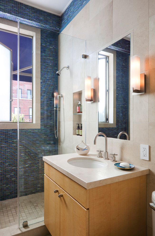 Modelo de cuarto de baño actual con armarios con paneles lisos, puertas de armario de madera clara, ducha esquinera, baldosas y/o azulejos azules, baldosas y/o azulejos en mosaico, aseo y ducha y lavabo bajoencimera
