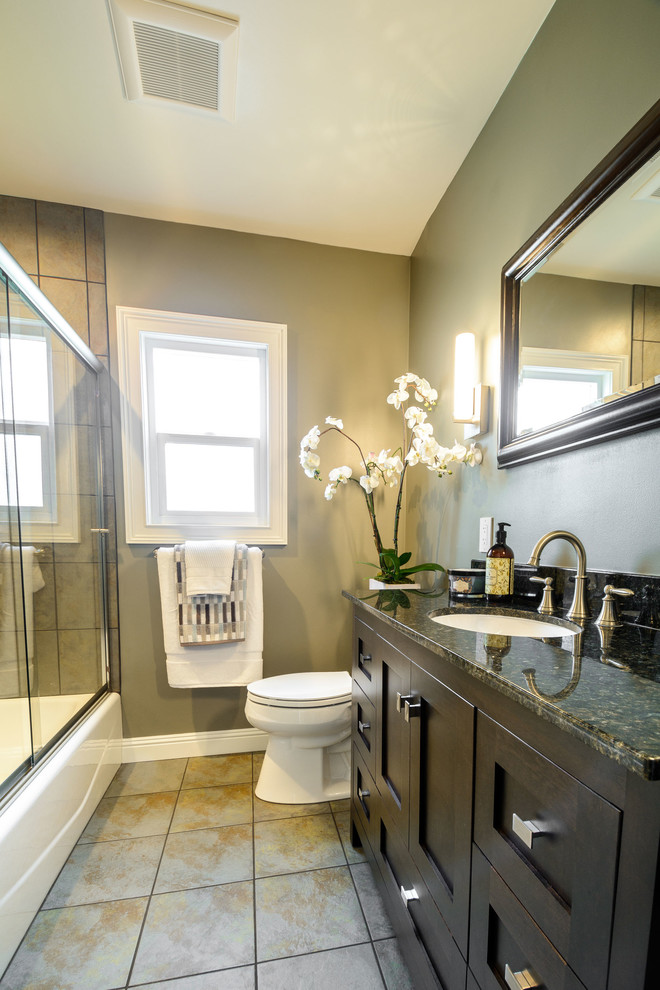 Bild på ett mellanstort funkis badrum, med skåp i shakerstil, bruna skåp, ett badkar i en alkov, en dusch/badkar-kombination, en toalettstol med separat cisternkåpa, beige kakel, porslinskakel, grå väggar, klinkergolv i porslin, ett undermonterad handfat och granitbänkskiva