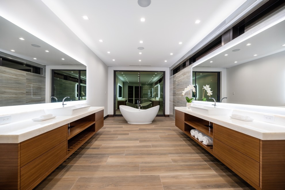 Идея дизайна: большая главная ванная комната в современном стиле с плоскими фасадами, фасадами цвета дерева среднего тона, отдельно стоящей ванной, серой плиткой, белыми стенами, настольной раковиной, коричневым полом и белой столешницей
